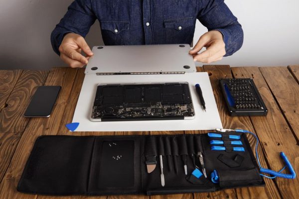 Service man opens backside topcase cover of computer laptop before repairing, cleaning and fixing it with his professional tools from toolkit box near on wooden table front view